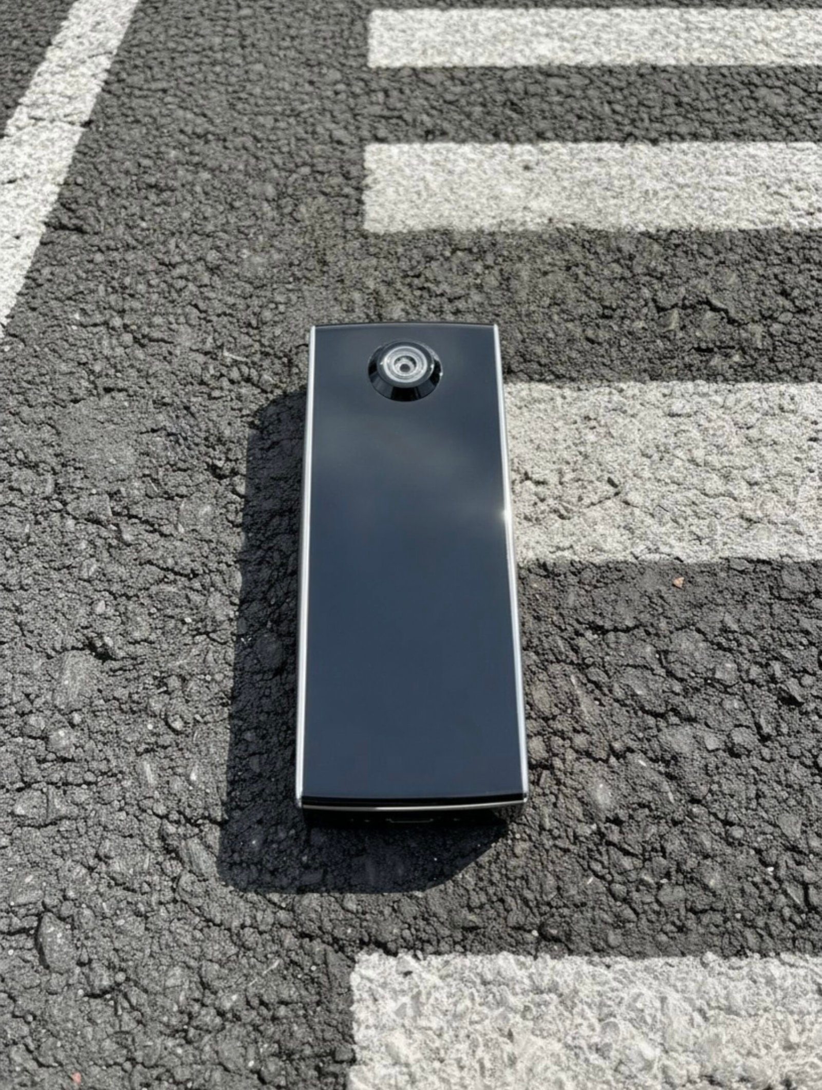 Alcometric X10 - front - Close-up of a black rectangular electronic device with a circular camera-like lens on rough asphalt next to a white crosswalk line.